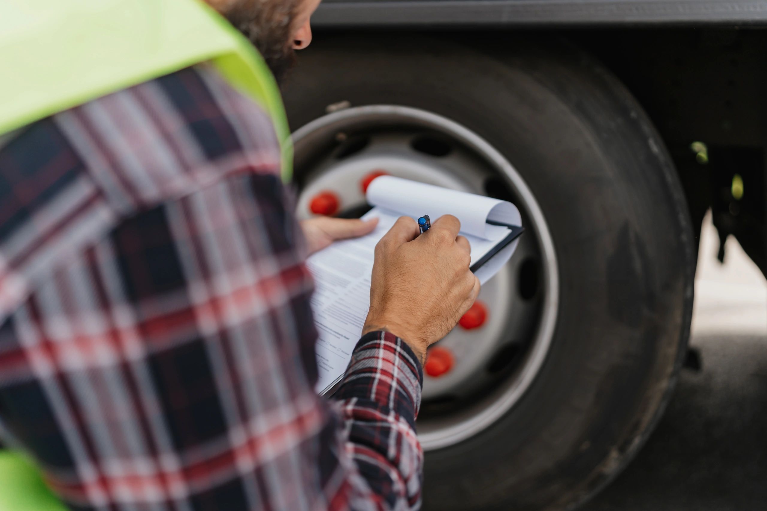 A black worker inspects a truck tire and fills out a checklist on a clipboard.
